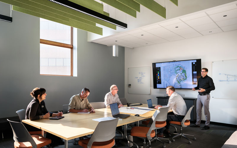 A man standing adjacent to a projection screen talking to a group of four at a conference table.