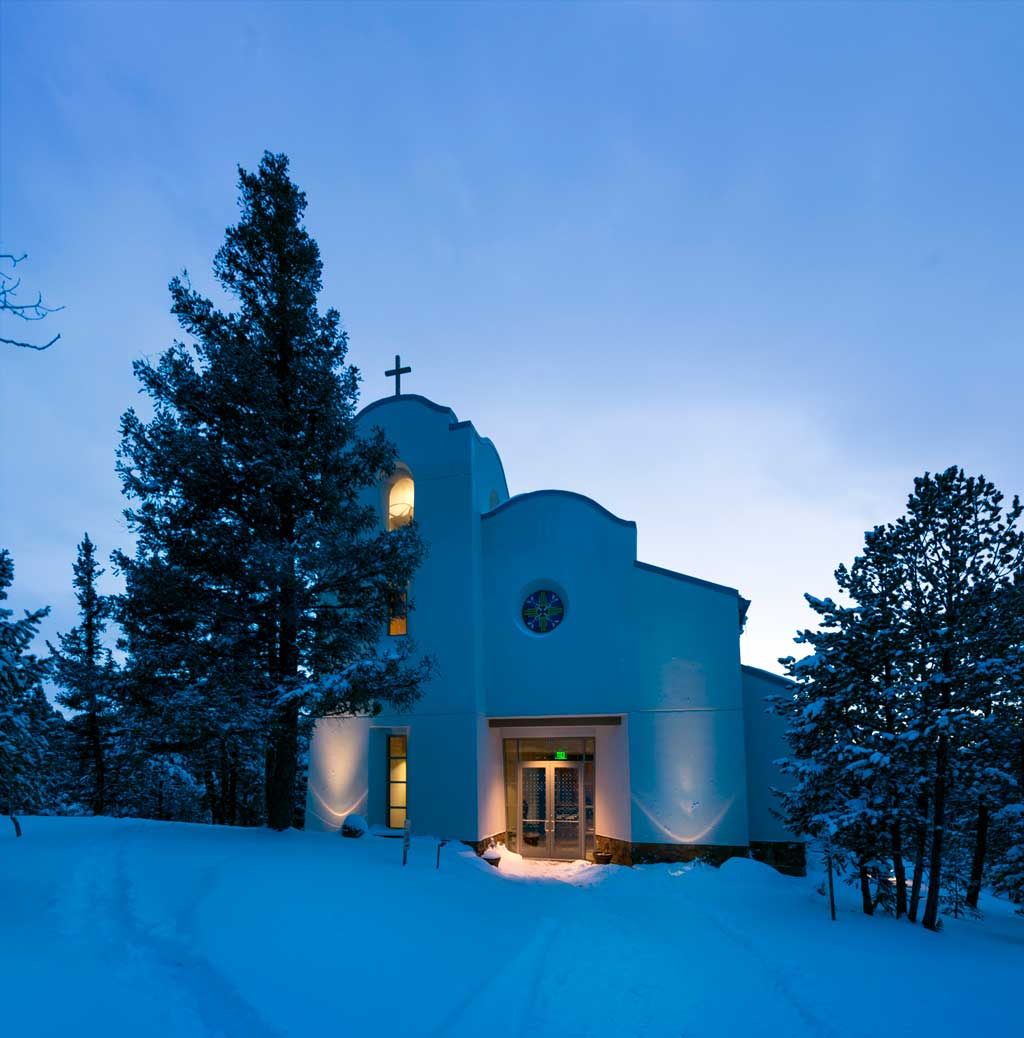 Nighttime exterior view of the Chapel at El Tesoro, illuminated by soft, warm lighting. The building features a modern yet rustic design with stone walls, large glass windows, and a welcoming entryway surrounded by a tranquil natural setting.