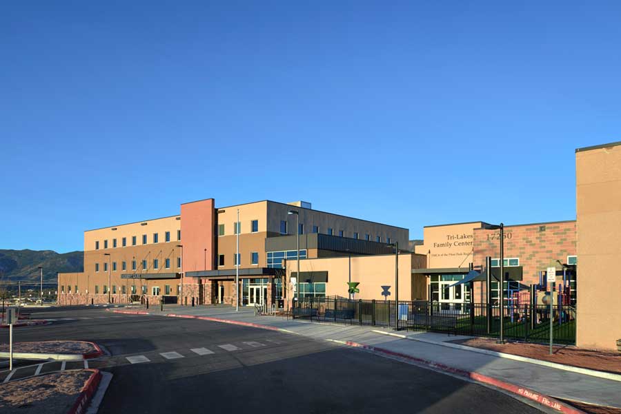 Exterior view of Tri-Lakes Community Health Pavilion, showcasing a contemporary design with a combination of glass, stone, and metal elements. The building is surrounded by well-maintained landscaping under a partly cloudy sky.