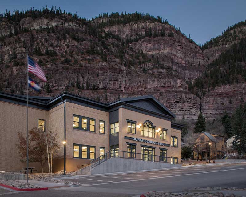 Exterior view of Ouray PK-12 School at dusk, with the building softly illuminated under the evening sky. The modern design features a mix of materials, large windows, and a welcoming entrance, creating an inviting atmosphere for students and visitors.