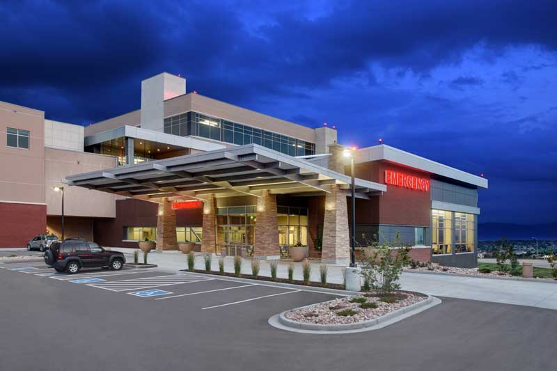 Nighttime exterior view of the SFMC IMCU/CCU expansion, showcasing a modern hospital building with large glass windows, clean architectural lines, and a combination of brick and metal finishes. The surrounding area is well-landscaped under a bright blue sky.