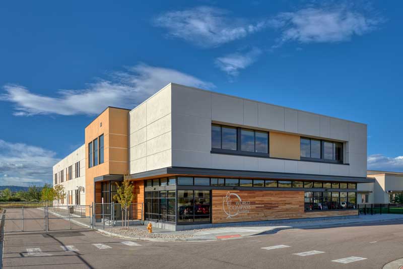 Exterior view of World Compass Charter School, showcasing a contemporary design with a combination of brick, metal, and large windows. The building is set against a bright sky, with a well-maintained landscape enhancing its welcoming appearance.