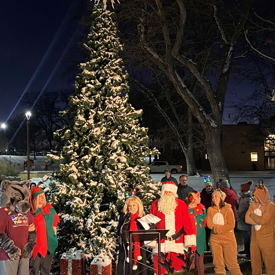 A photo of a group of adults wearing coats, hats, and gloves; adults in Santa, reindeer, and elf costumes; and an adult in the Pueblo Community College mascot panther costume listen to a speech by a representative from Pueblo Community College in front of a decorated Christmas tree on a snowy cold night.