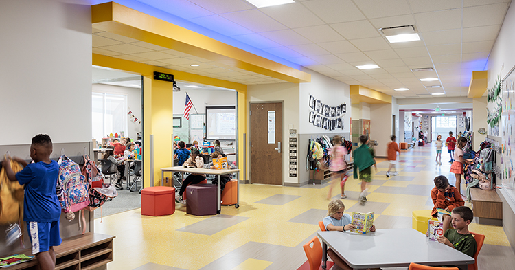 a bright and colorful hallway in woodland elementary school. one student is hanging up a backpack, two students sit at a table in the hall, several students are walking down the hall, and a classroom full of students can be seen through two large doorways