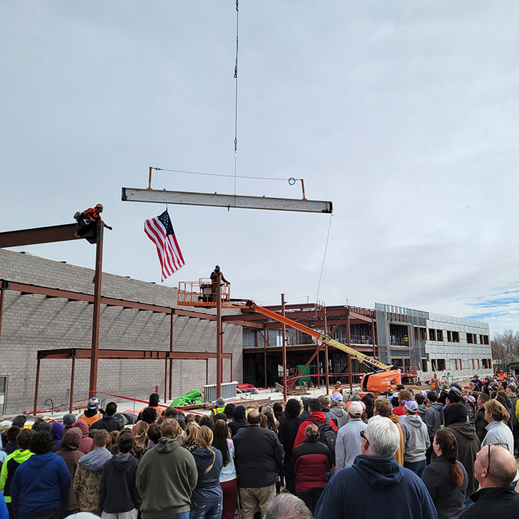 a crowd of school children and adults watch a last structural beam being lowered into place with a crane