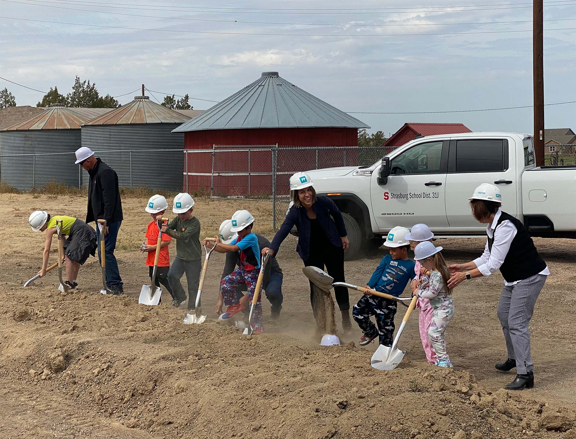 students and adults wearing hard hats participate in the groundbreaking by ceremoniously shoveling dirt